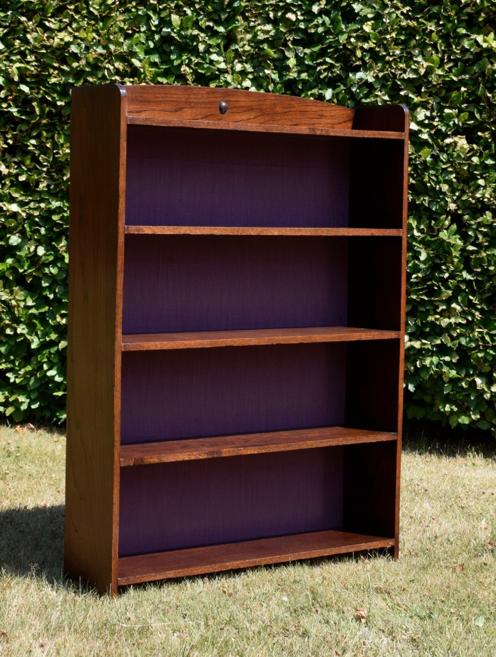 An Arts and Crafts solid oak open bookcase featuring five shelves, solid plank sides, an arched gallery top with a central decorative motif, and a purple fabric-lined back panel. View XI