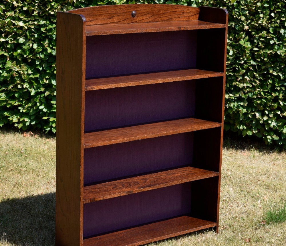 An Arts and Crafts solid oak open bookcase featuring five shelves, solid plank sides, an arched gallery top with a central decorative motif, and a purple fabric-lined back panel. View XII