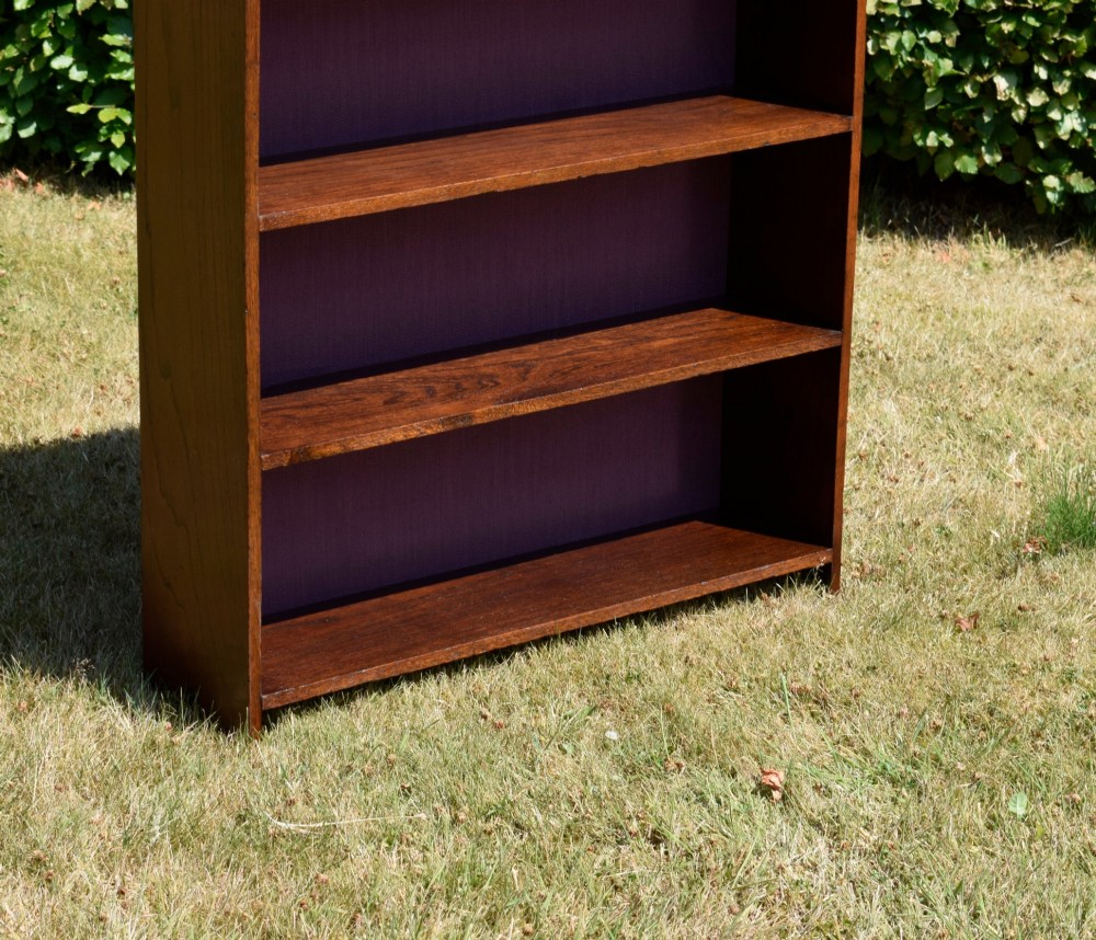An Arts and Crafts solid oak open bookcase featuring five shelves, solid plank sides, an arched gallery top with a central decorative motif, and a purple fabric-lined back panel. View XIII