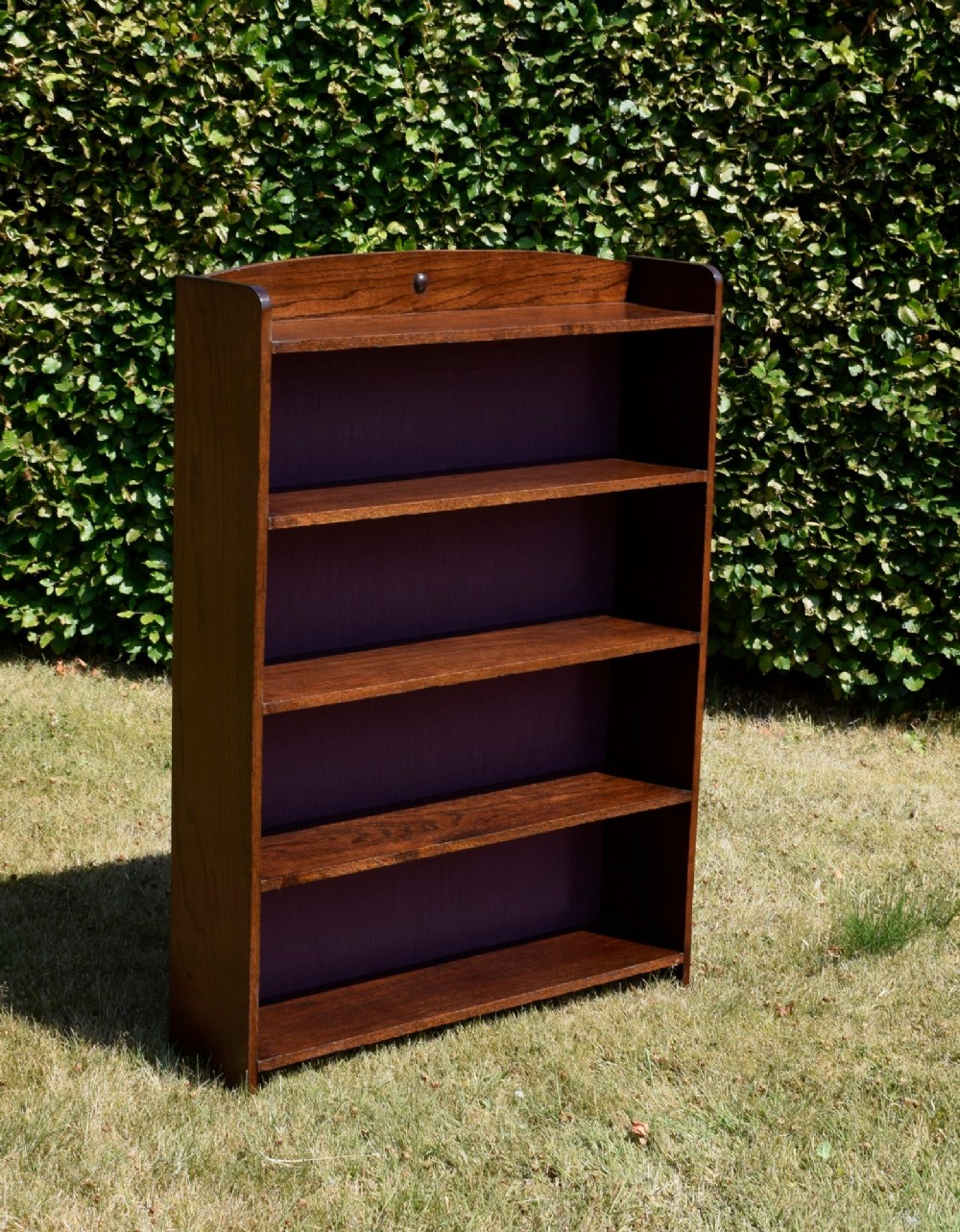 An Arts and Crafts solid oak open bookcase featuring five shelves, solid plank sides, an arched gallery top with a central decorative motif, and a purple fabric-lined back panel. View XIV