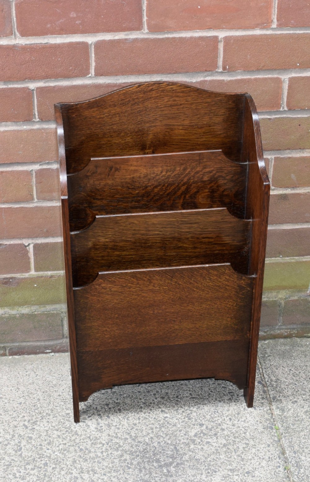 A dark oak Arts and Crafts freestanding magazine rack featuring a gently arched crest rail and three tiered open compartments.