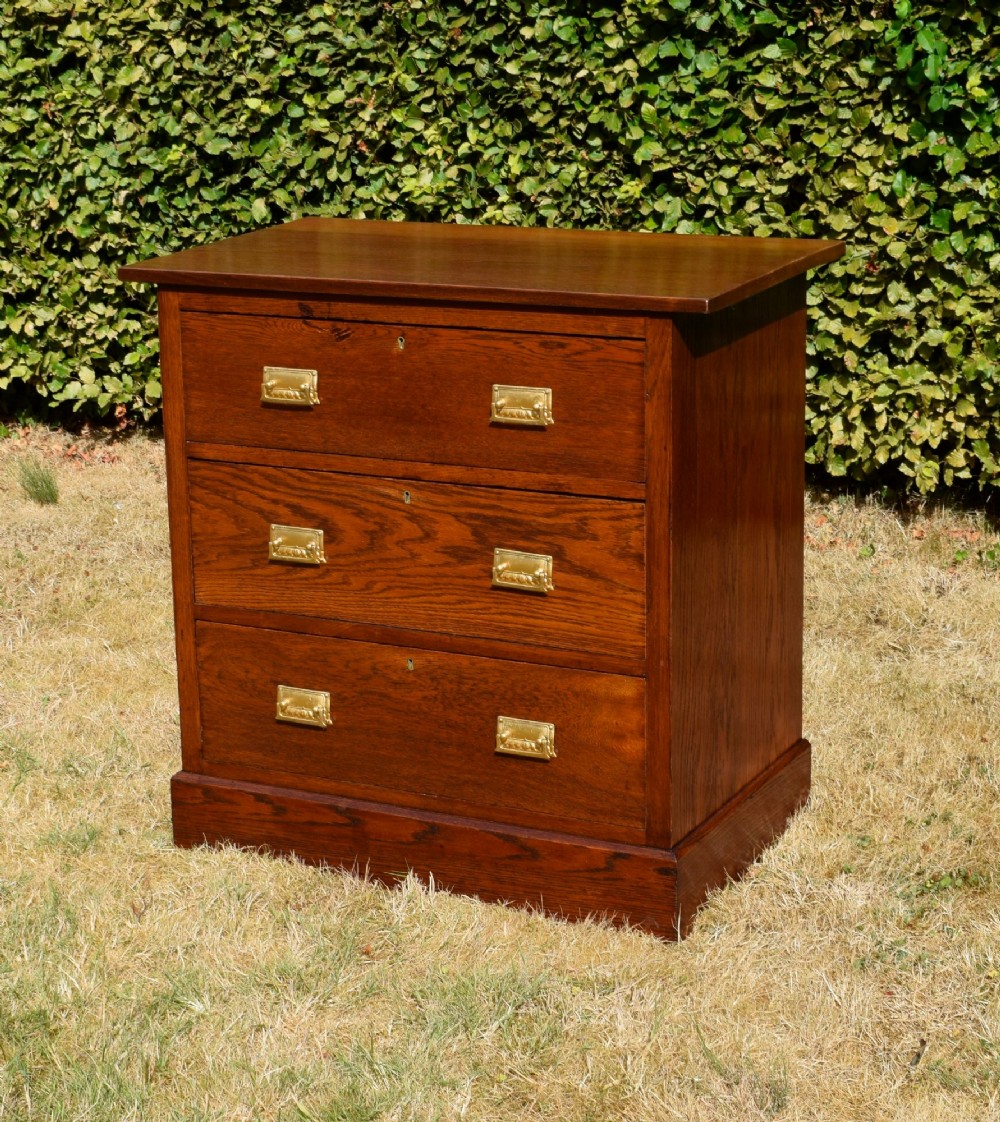A medium-toned oak chest of drawers featuring three drawers with rectangular brass handles resting on a plinth base. View II