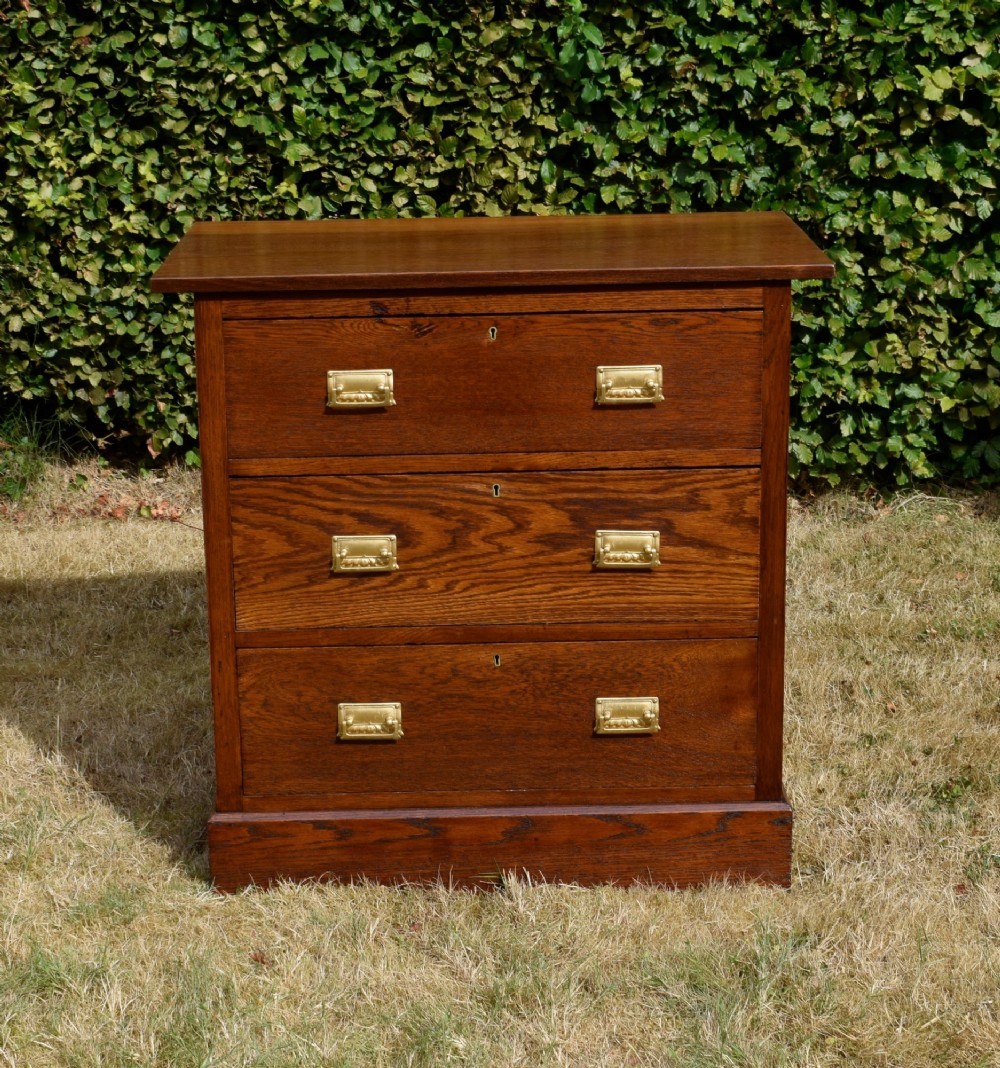 A medium-toned oak chest of drawers featuring three drawers with rectangular brass handles resting on a plinth base. View III