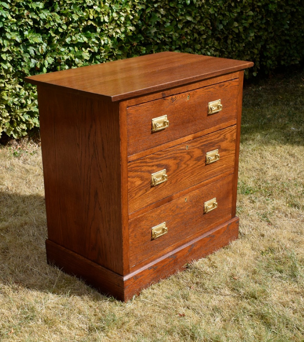 A medium-toned oak chest of drawers featuring three drawers with rectangular brass handles resting on a plinth base. View IV