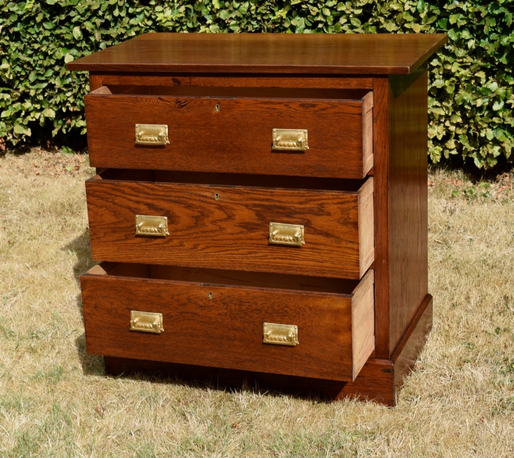 A medium-toned oak chest of drawers featuring three drawers with rectangular brass handles resting on a plinth base. View V
