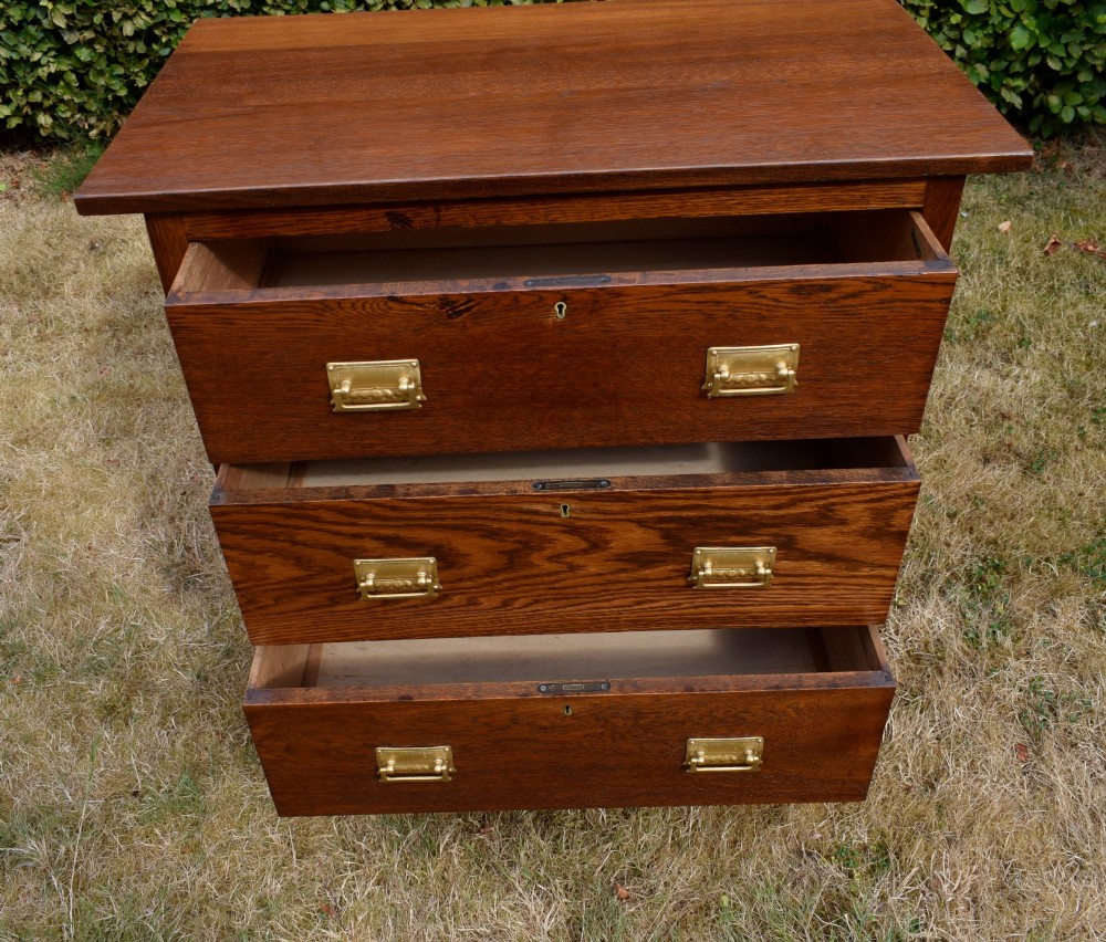A medium-toned oak chest of drawers featuring three drawers with rectangular brass handles resting on a plinth base. View VI