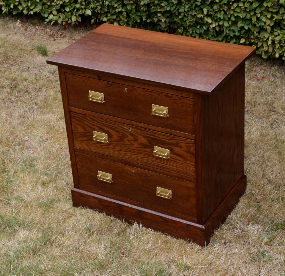 A medium-toned oak chest of drawers featuring three drawers with rectangular brass handles resting on a plinth base. View VIII