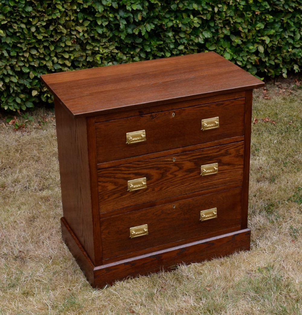 A medium-toned oak chest of drawers featuring three drawers with rectangular brass handles resting on a plinth base. View IX