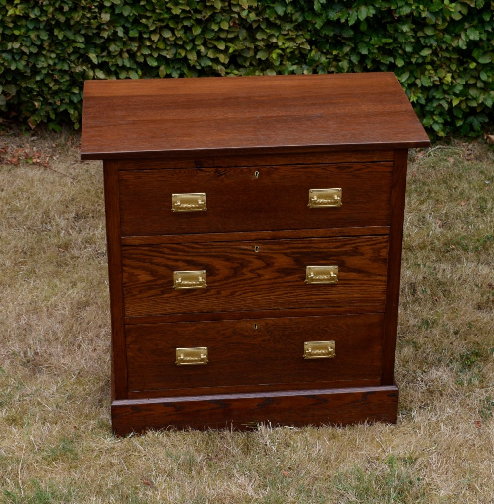 A medium-toned oak chest of drawers featuring three drawers with rectangular brass handles resting on a plinth base. View X