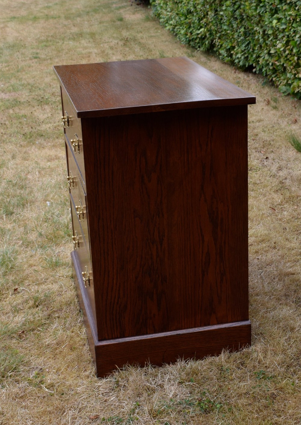 A medium-toned oak chest of drawers featuring three drawers with rectangular brass handles resting on a plinth base. View XV