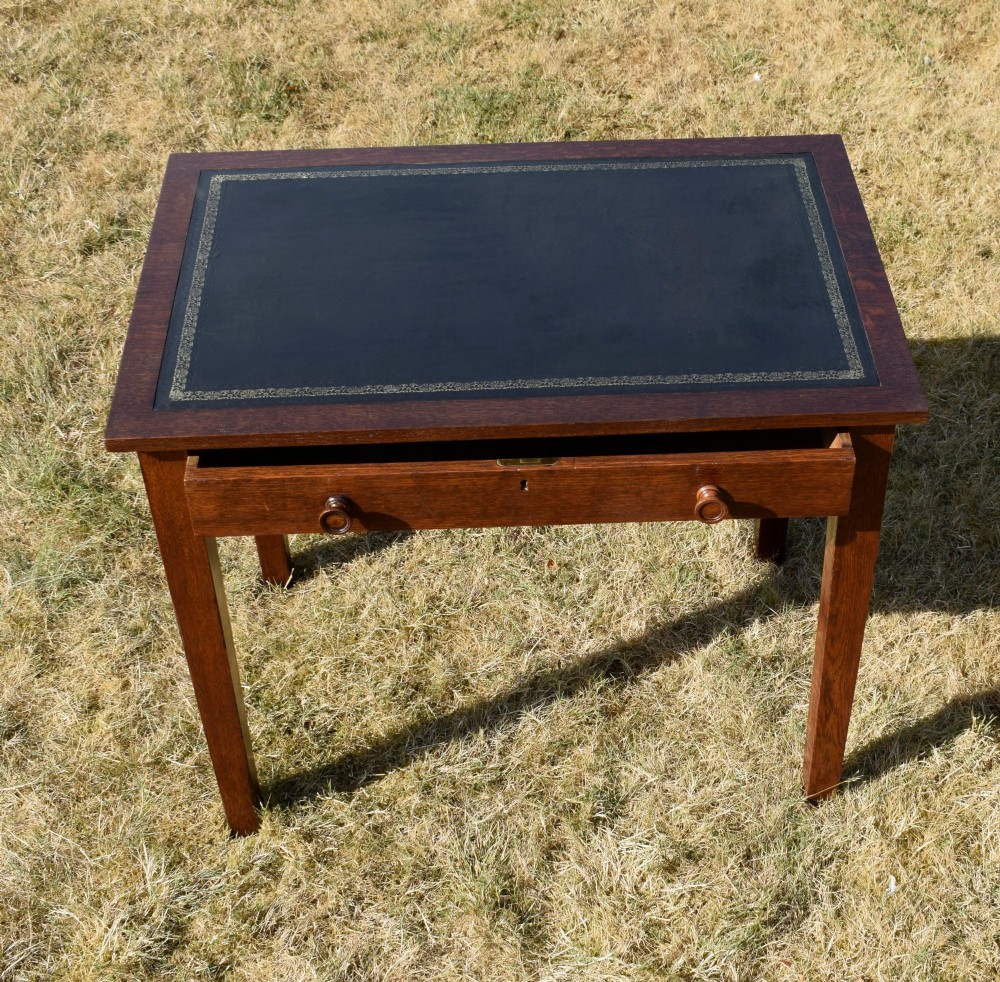 A warm brown oak Arts and Crafts writing desk with a dark inset leather top, a single drawer with turned wooden knobs, and four square straight legs. View V