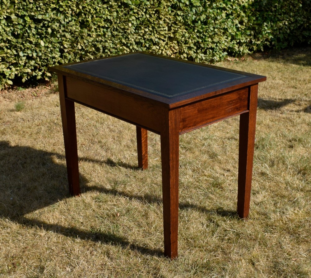 A warm brown oak Arts and Crafts writing desk with a dark inset leather top, a single drawer with turned wooden knobs, and four square straight legs. View VI