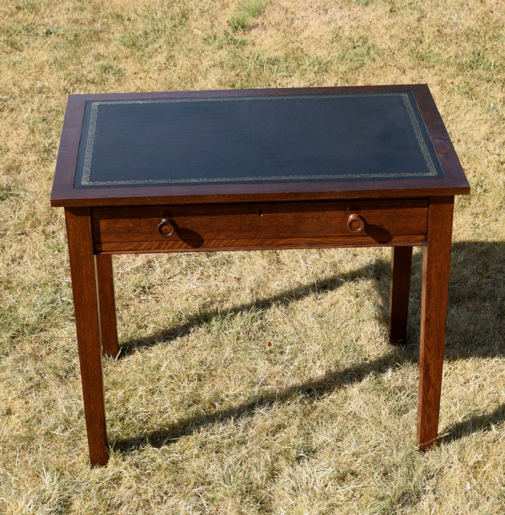 A warm brown oak Arts and Crafts writing desk with a dark inset leather top, a single drawer with turned wooden knobs, and four square straight legs. View VII