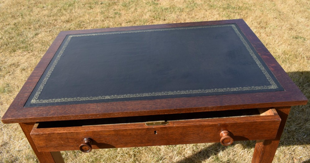 A warm brown oak Arts and Crafts writing desk with a dark inset leather top, a single drawer with turned wooden knobs, and four square straight legs. View VIII