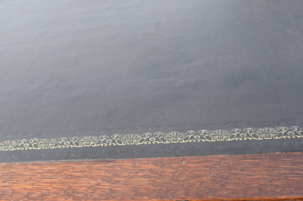A warm brown oak Arts and Crafts writing desk with a dark inset leather top, a single drawer with turned wooden knobs, and four square straight legs. View IX