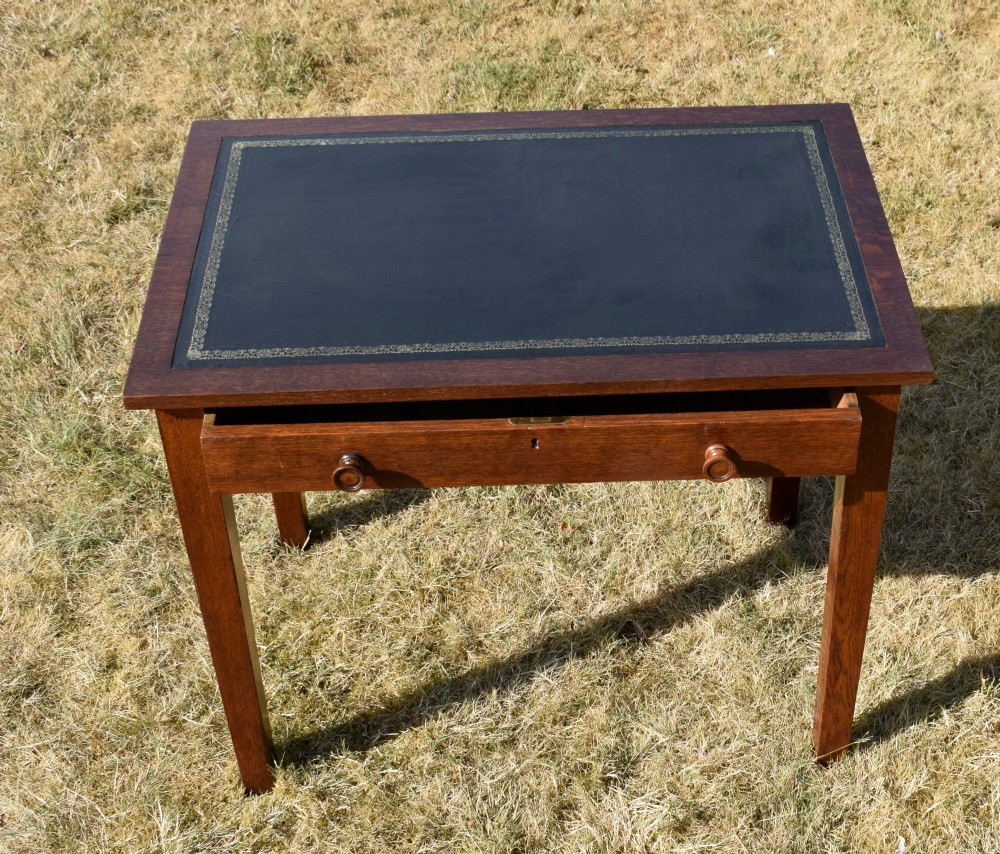 A warm brown oak Arts and Crafts writing desk with a dark inset leather top, a single drawer with turned wooden knobs, and four square straight legs. View XI