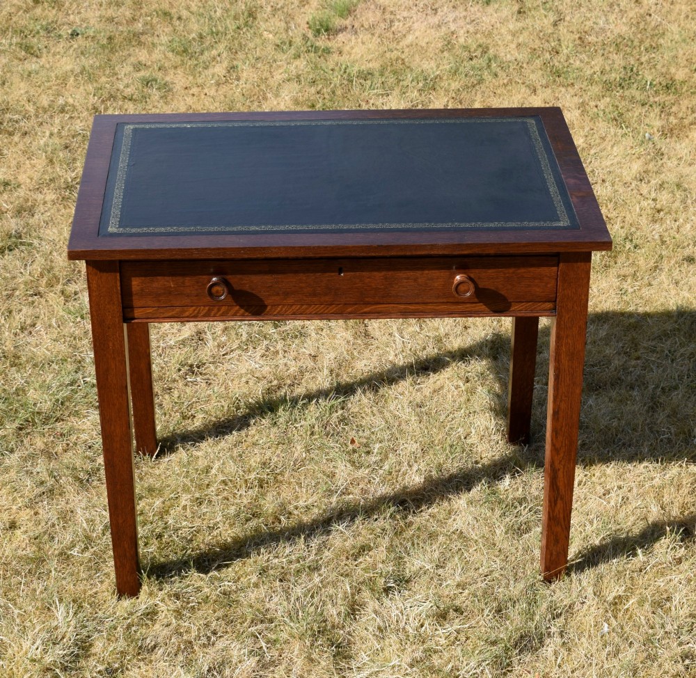 A warm brown oak Arts and Crafts writing desk with a dark inset leather top, a single drawer with turned wooden knobs, and four square straight legs. View XII