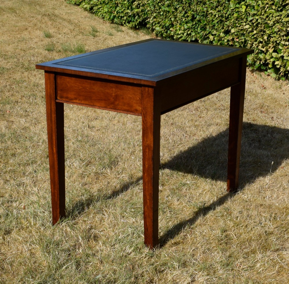A warm brown oak Arts and Crafts writing desk with a dark inset leather top, a single drawer with turned wooden knobs, and four square straight legs. View XIII