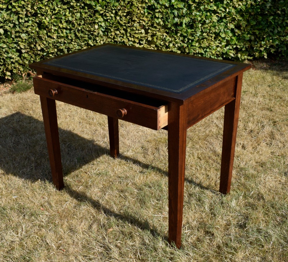 A warm brown oak Arts and Crafts writing desk with a dark inset leather top, a single drawer with turned wooden knobs, and four square straight legs. View XIV