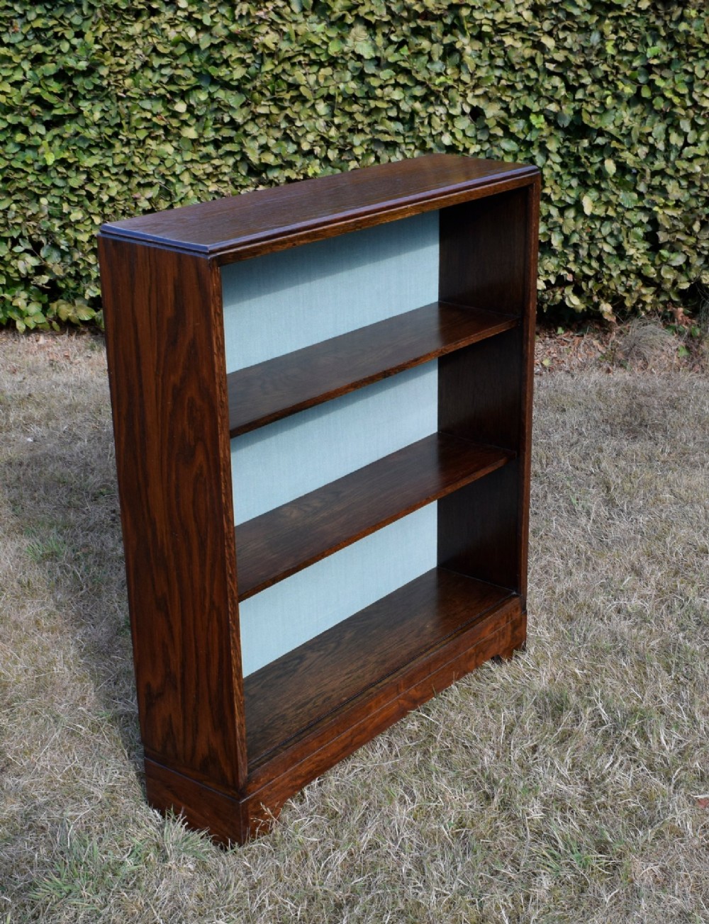 A dark oak Art Deco open bookcase featuring three shelves and a light duck-egg blue textured back panel. View II