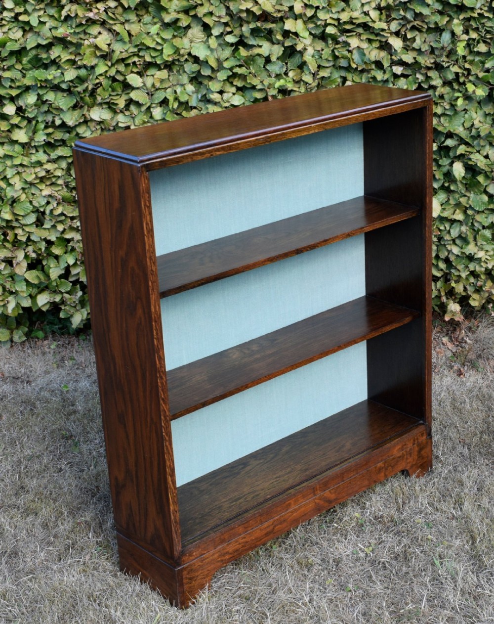 A dark oak Art Deco open bookcase featuring three shelves and a light duck-egg blue textured back panel. View XII