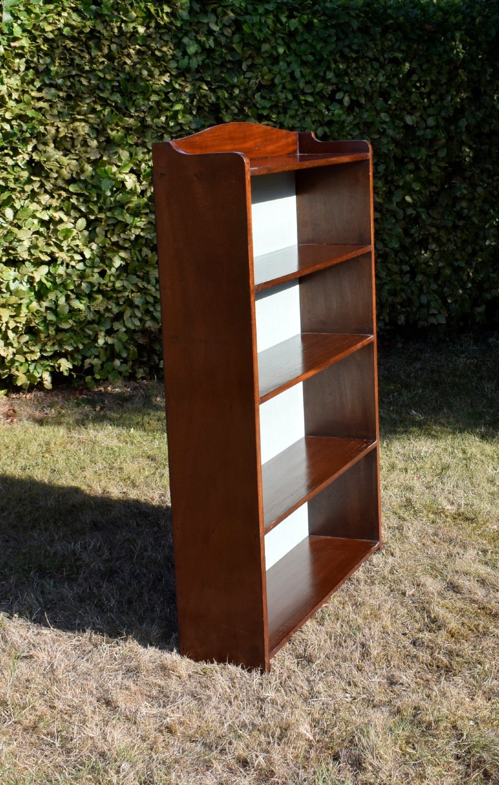 A standing mahogany open bookcase featuring four horizontal shelves, a gently curved top upstand, and a light-coloured fabric back panel. View V