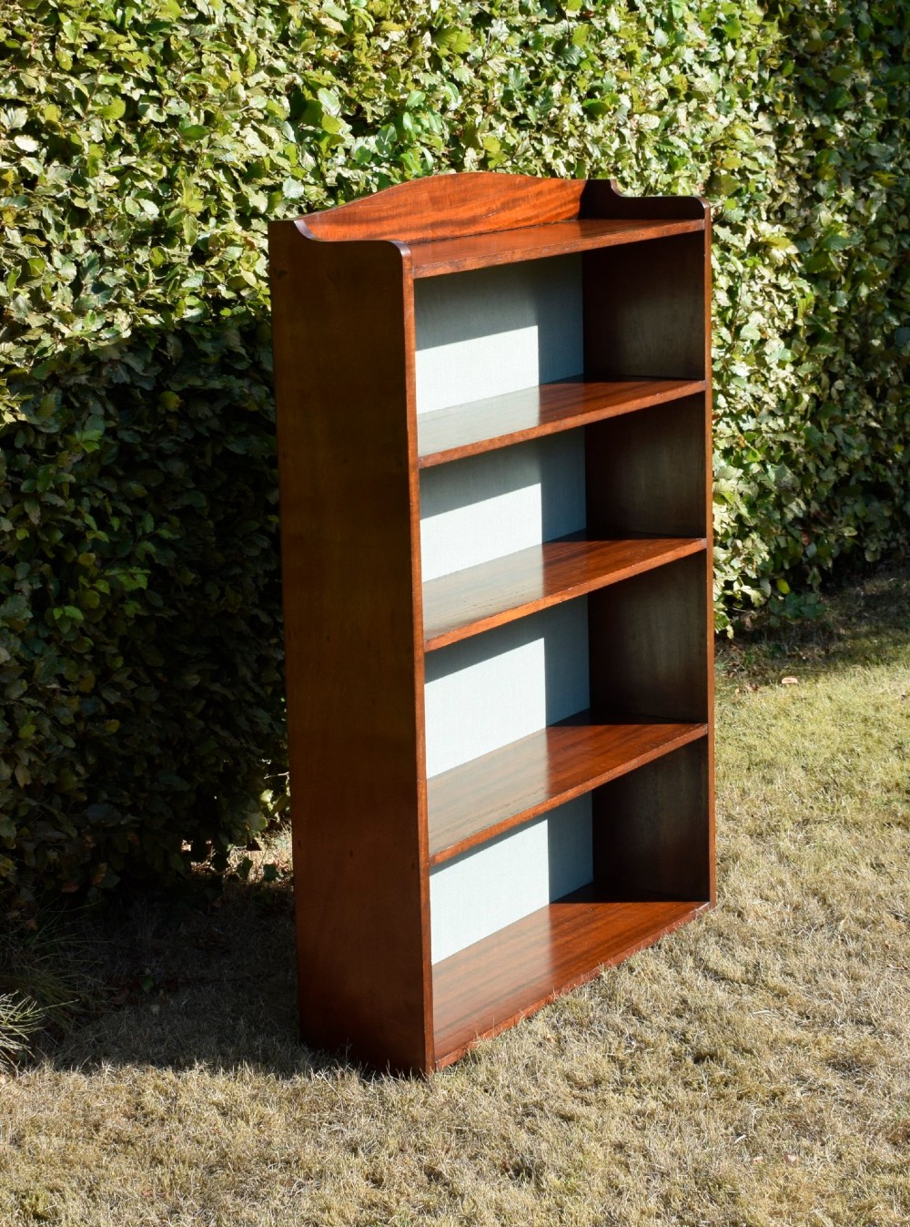 A standing mahogany open bookcase featuring four horizontal shelves, a gently curved top upstand, and a light-coloured fabric back panel. View IX