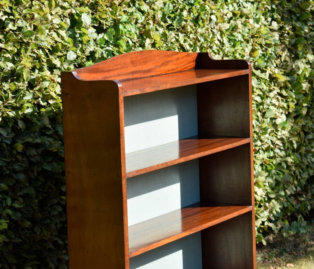 A standing mahogany open bookcase featuring four horizontal shelves, a gently curved top upstand, and a light-coloured fabric back panel. View X