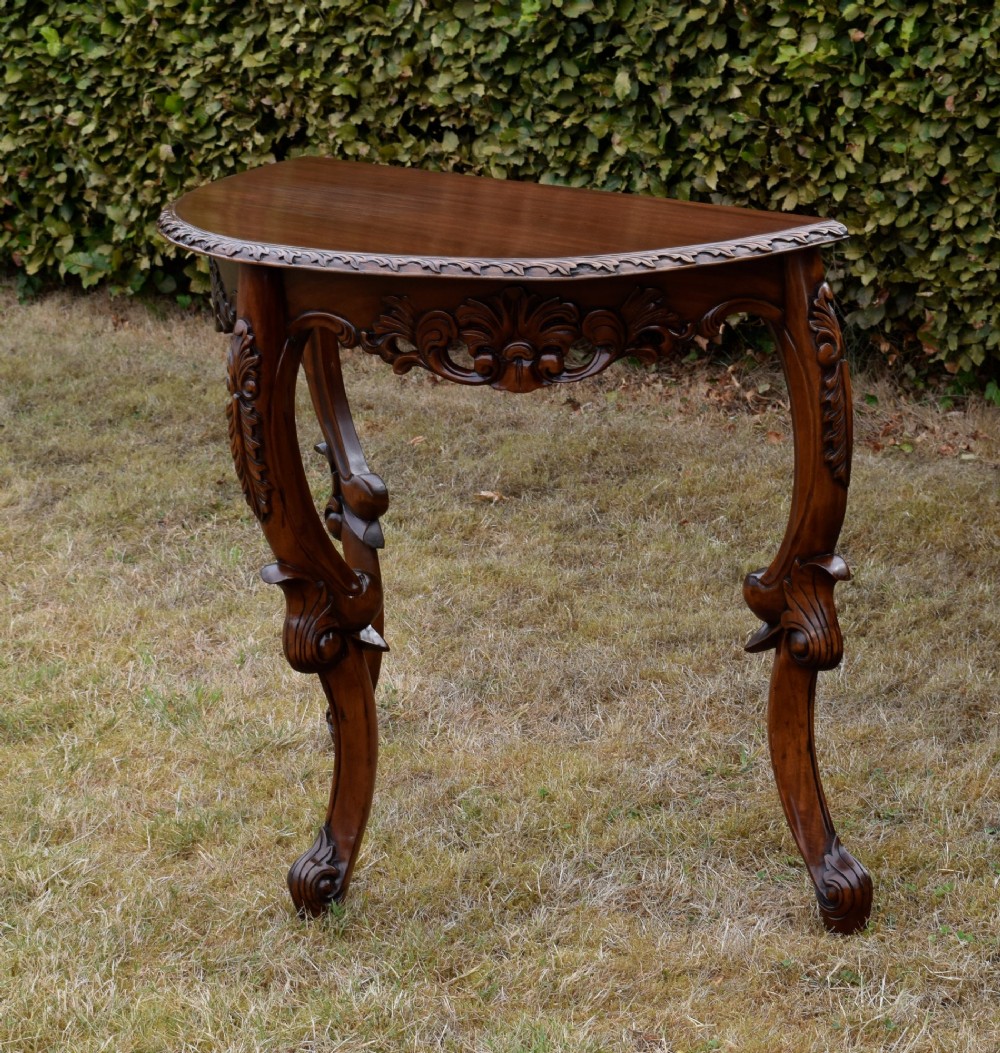 A dark mahogany demi-lune console table featuring deep acanthus leaf carvings on the frieze and three cabriole legs. View V
