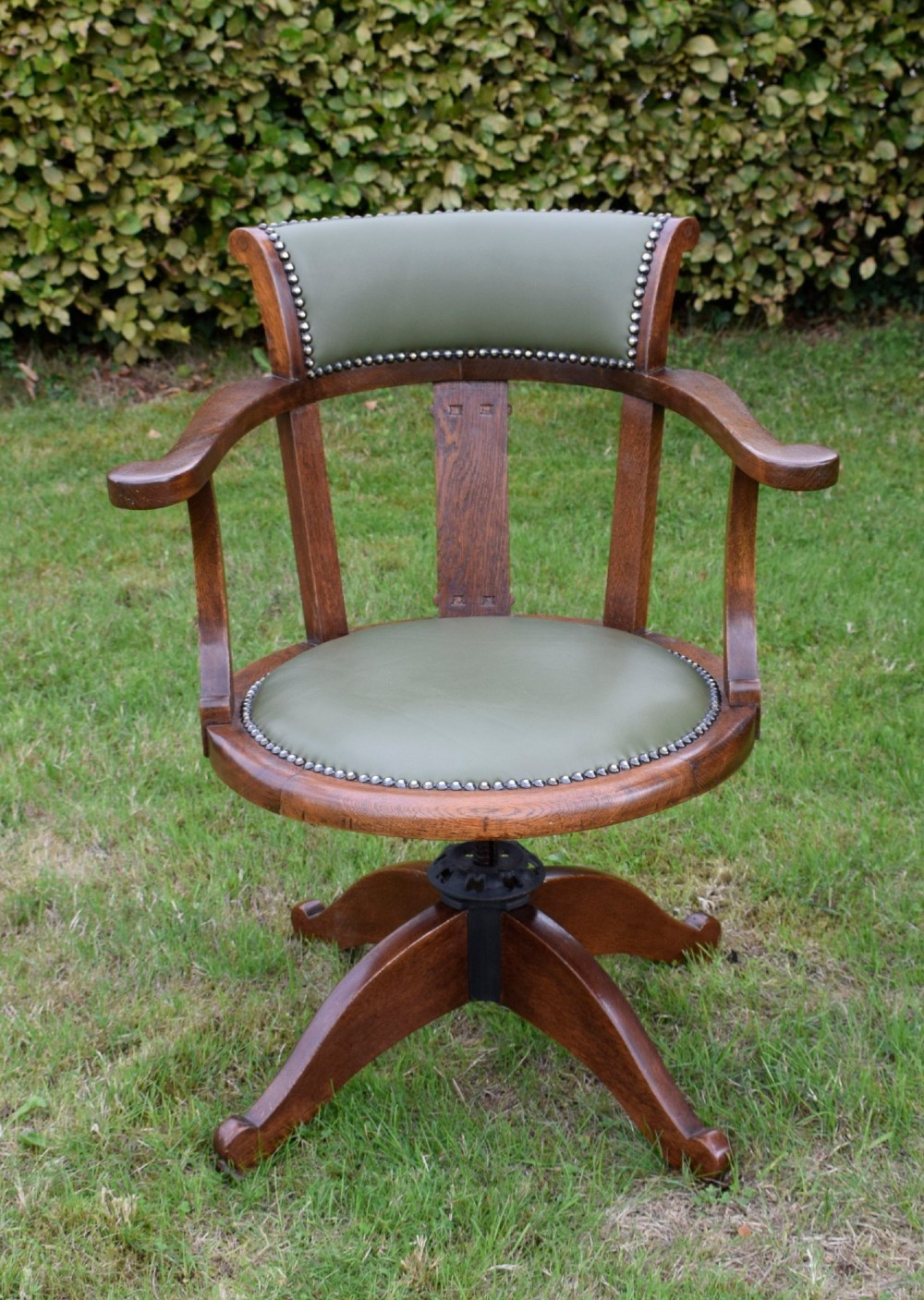 An early 20th-century oak swivel desk chair featuring a green leather padded seat and backrest trimmed with metal studs, supported by a four-spoke wooden base with a central iron mechanism. View II