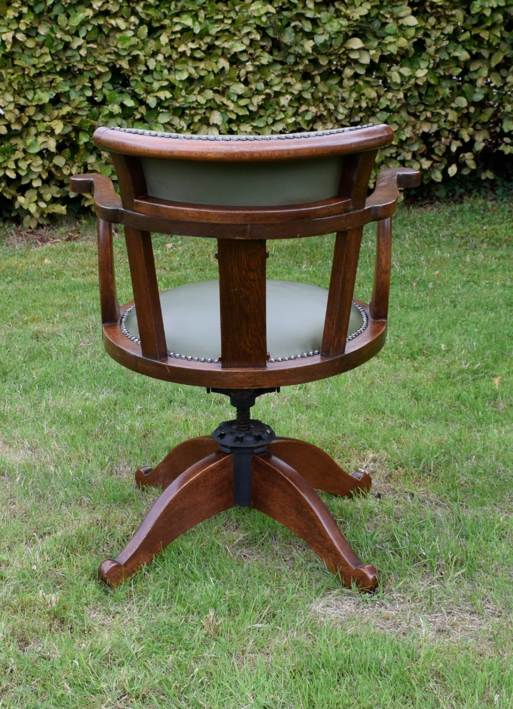 An early 20th-century oak swivel desk chair featuring a green leather padded seat and backrest trimmed with metal studs, supported by a four-spoke wooden base with a central iron mechanism. View V