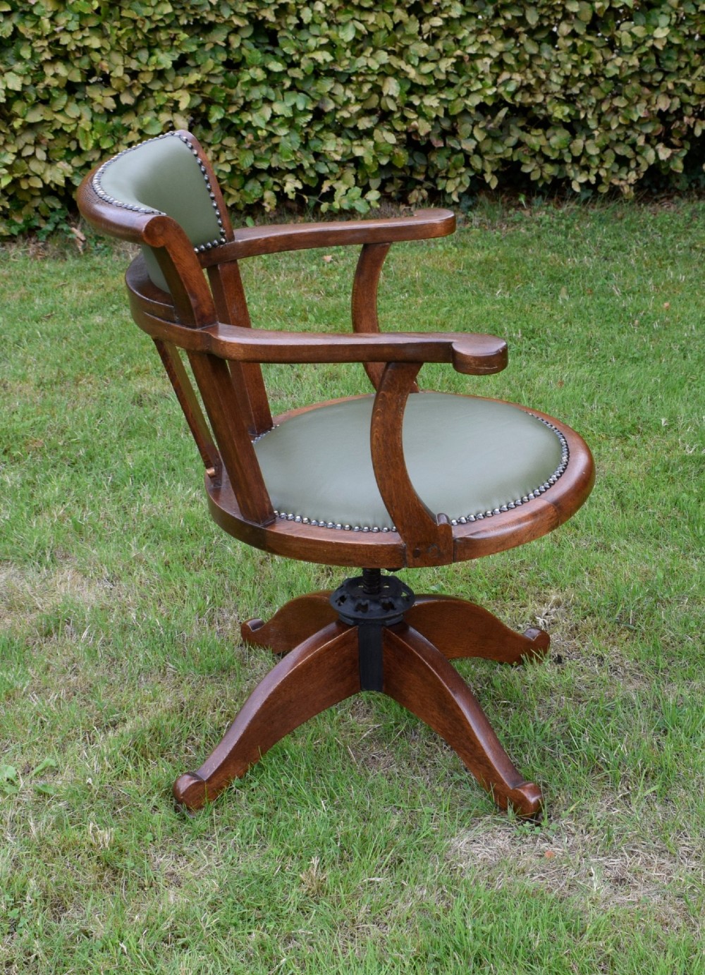An early 20th-century oak swivel desk chair featuring a green leather padded seat and backrest trimmed with metal studs, supported by a four-spoke wooden base with a central iron mechanism. View VI
