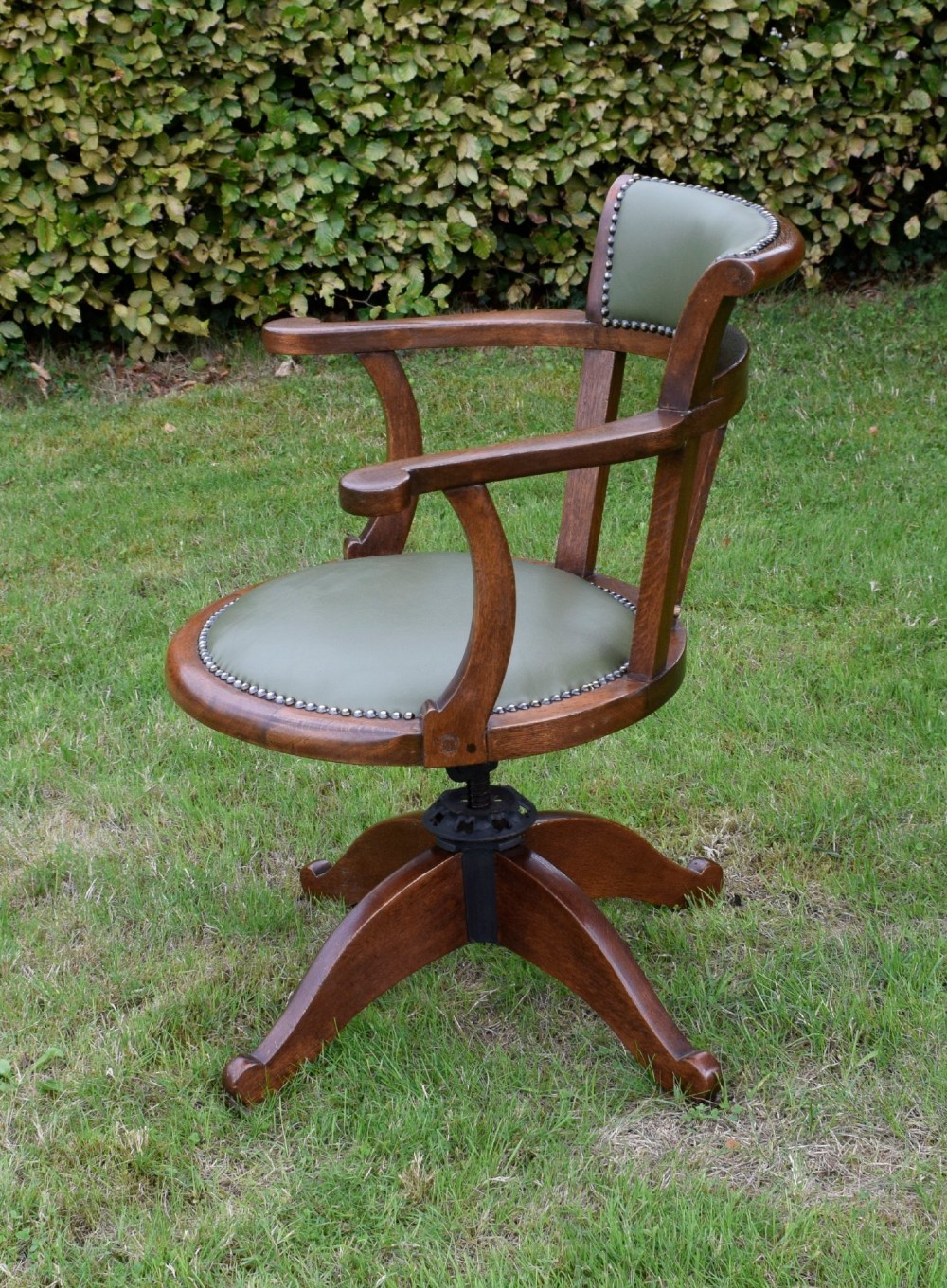 An early 20th-century oak swivel desk chair featuring a green leather padded seat and backrest trimmed with metal studs, supported by a four-spoke wooden base with a central iron mechanism. View VII
