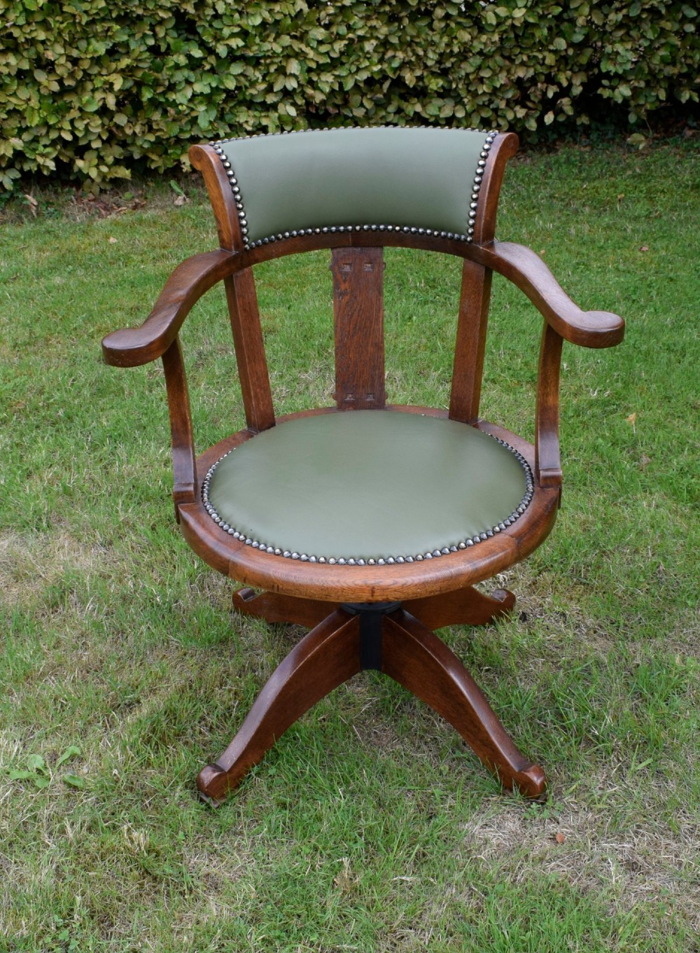 An early 20th-century oak swivel desk chair featuring a green leather padded seat and backrest trimmed with metal studs, supported by a four-spoke wooden base with a central iron mechanism. View IX