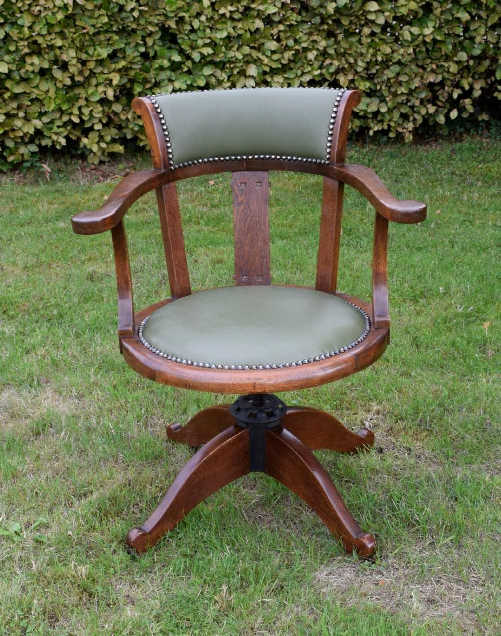 An early 20th-century oak swivel desk chair featuring a green leather padded seat and backrest trimmed with metal studs, supported by a four-spoke wooden base with a central iron mechanism. View X
