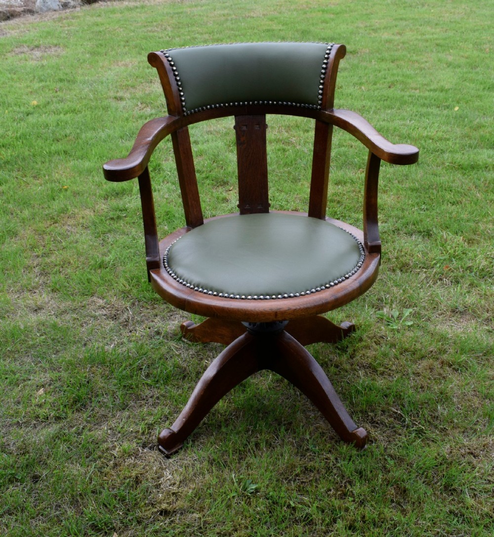An early 20th-century oak swivel desk chair featuring a green leather padded seat and backrest trimmed with metal studs, supported by a four-spoke wooden base with a central iron mechanism. View XIII