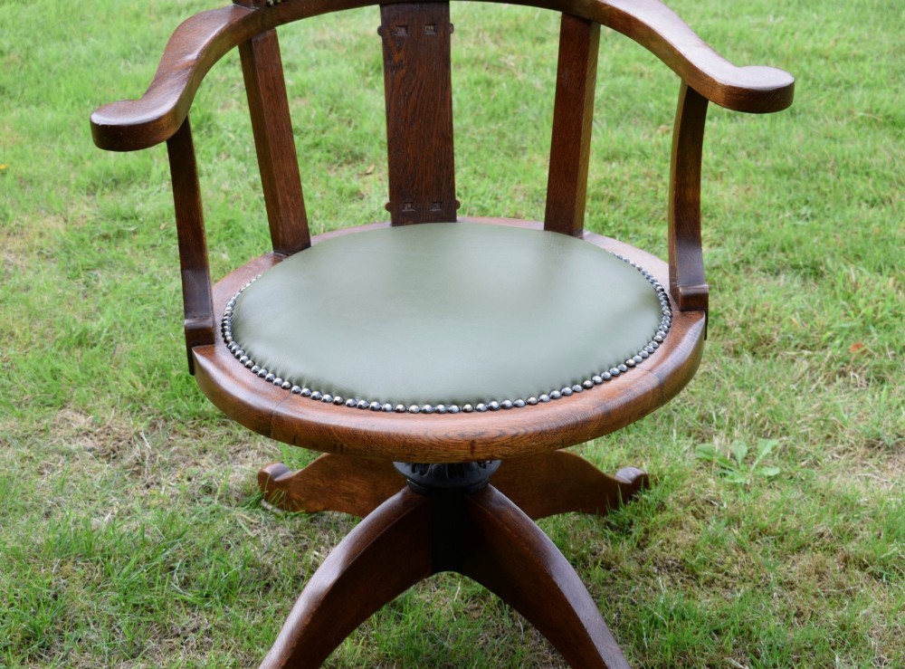 An early 20th-century oak swivel desk chair featuring a green leather padded seat and backrest trimmed with metal studs, supported by a four-spoke wooden base with a central iron mechanism. View XIV