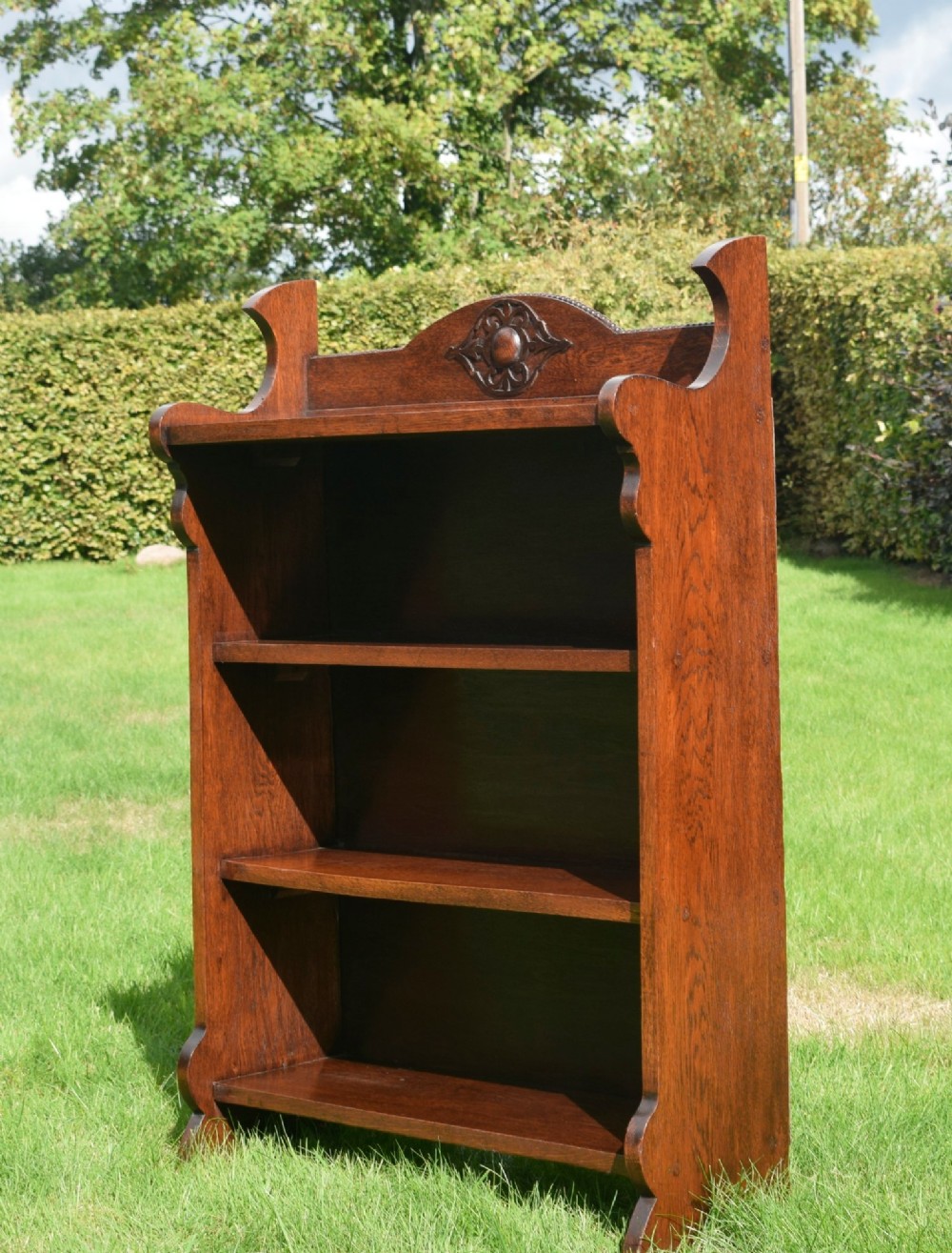An early twentieth-century open oak bookcase featuring a carved floral crest on the top gallery, shaped sides, and three fixed shelves.