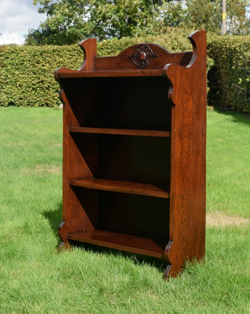 An early twentieth-century open oak bookcase featuring a carved floral crest on the top gallery, shaped sides, and three fixed shelves. View II