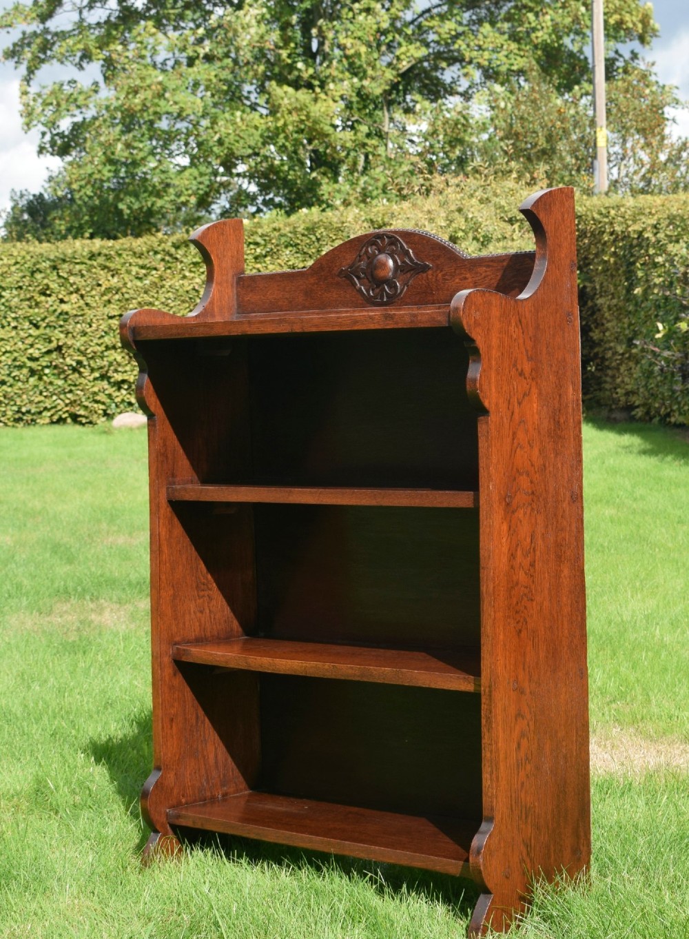An early twentieth-century open oak bookcase featuring a carved floral crest on the top gallery, shaped sides, and three fixed shelves. View IV