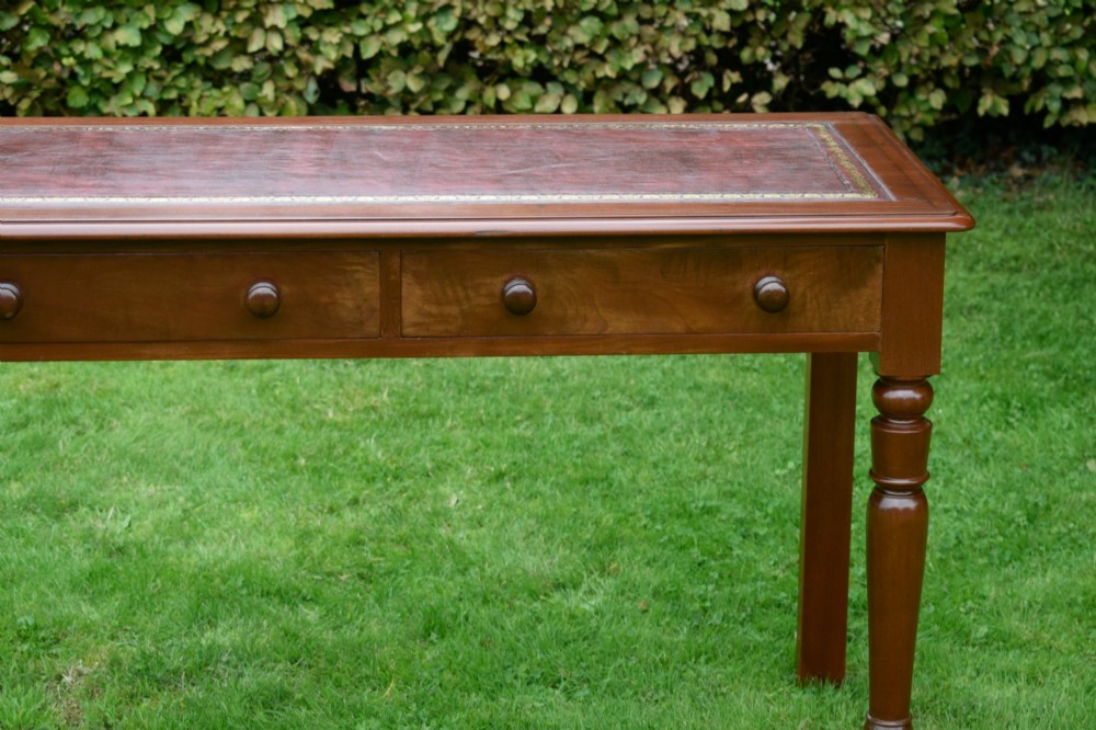 A Victorian mahogany writing table with a red leather inset surface, two drawers with wooden knob handles, turned front legs, and square back legs. View VI