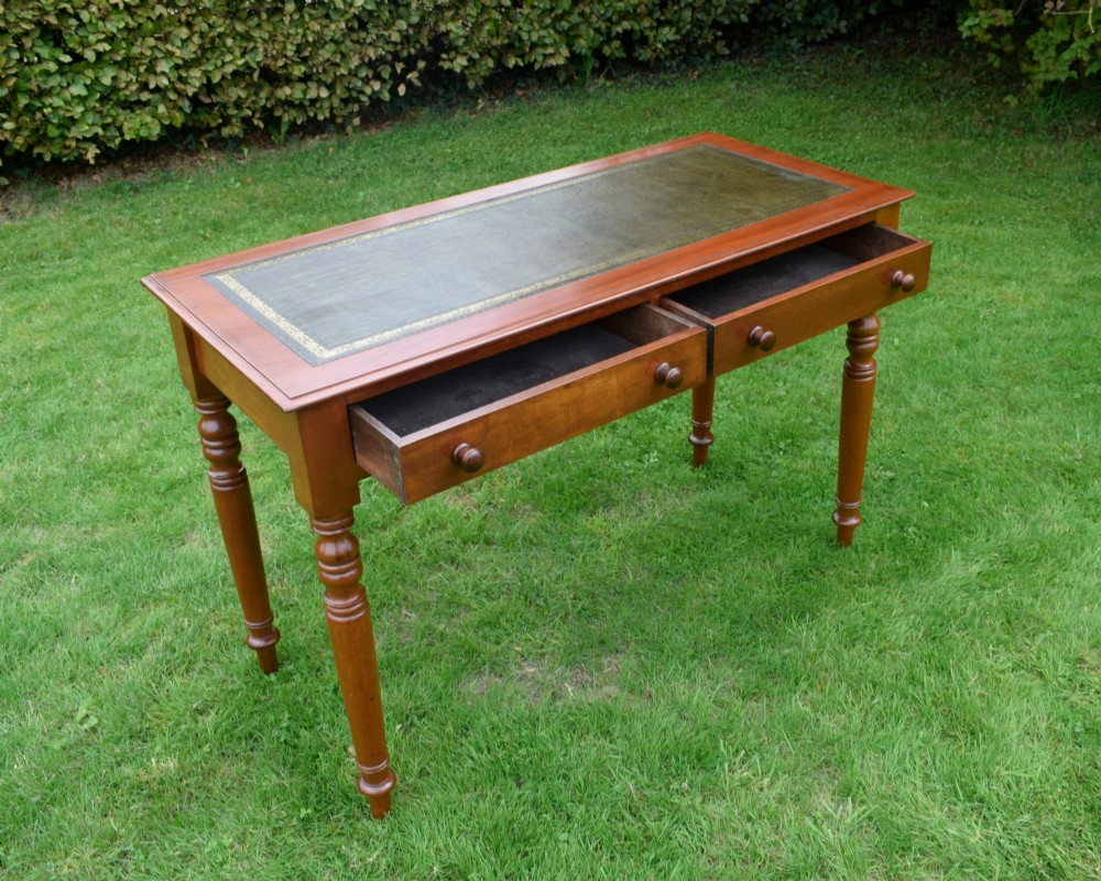 A Victorian warm brown mahogany writing table featuring an inset green leather writing surface, two drawers with knob handles, and turned legs. View X