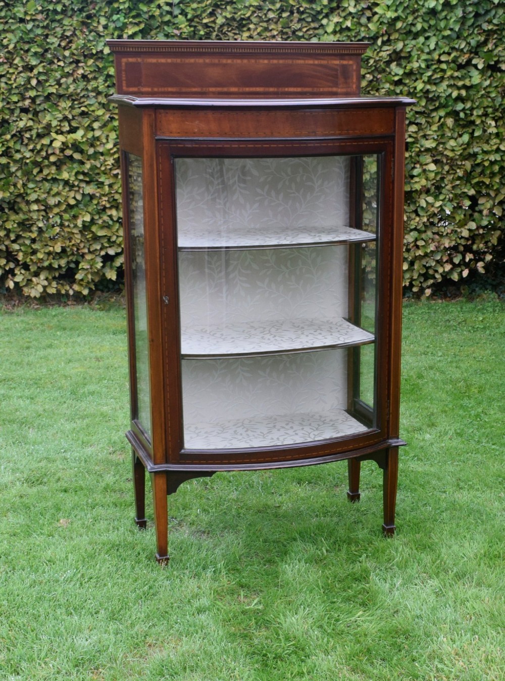 An Edwardian mahogany bowfront display cabinet featuring curved glass doors, satinwood string inlay, lined interior shelves, and tapered square legs. View III