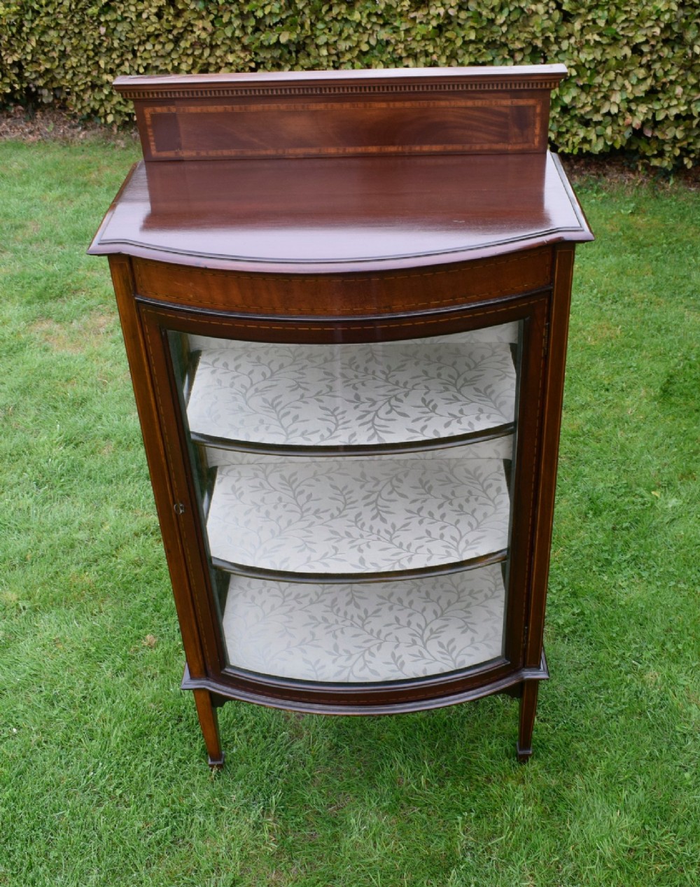 An Edwardian mahogany bowfront display cabinet featuring curved glass doors, satinwood string inlay, lined interior shelves, and tapered square legs. View VII