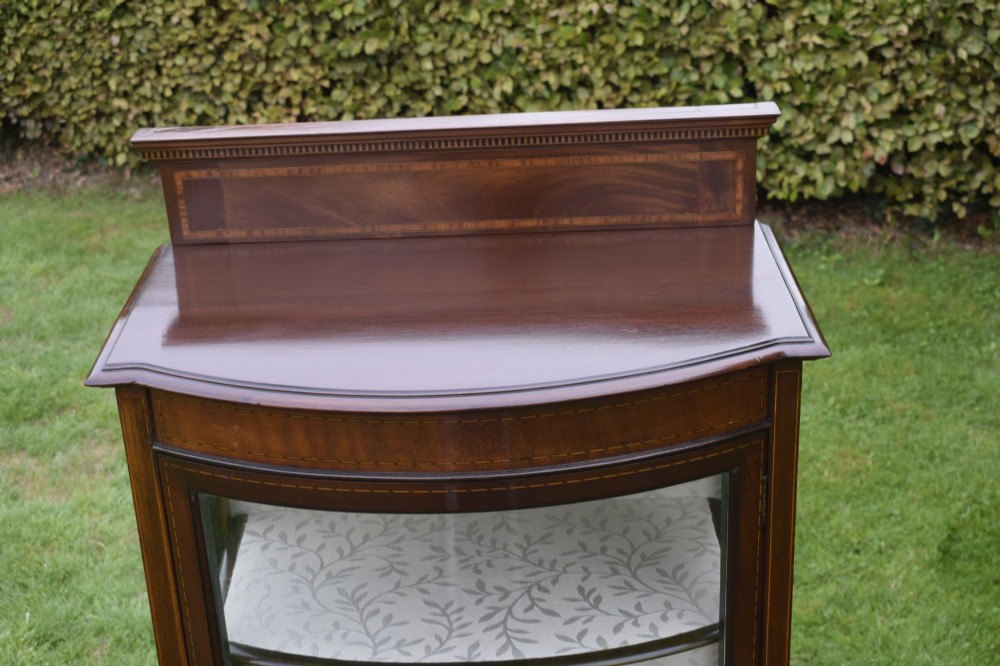 An Edwardian mahogany bowfront display cabinet featuring curved glass doors, satinwood string inlay, lined interior shelves, and tapered square legs. View VIII