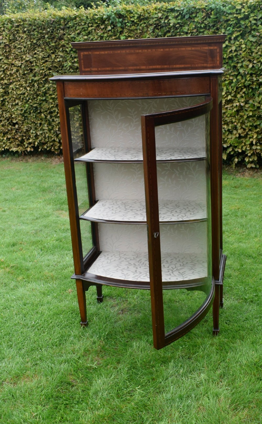 An Edwardian mahogany bowfront display cabinet featuring curved glass doors, satinwood string inlay, lined interior shelves, and tapered square legs. View XIV