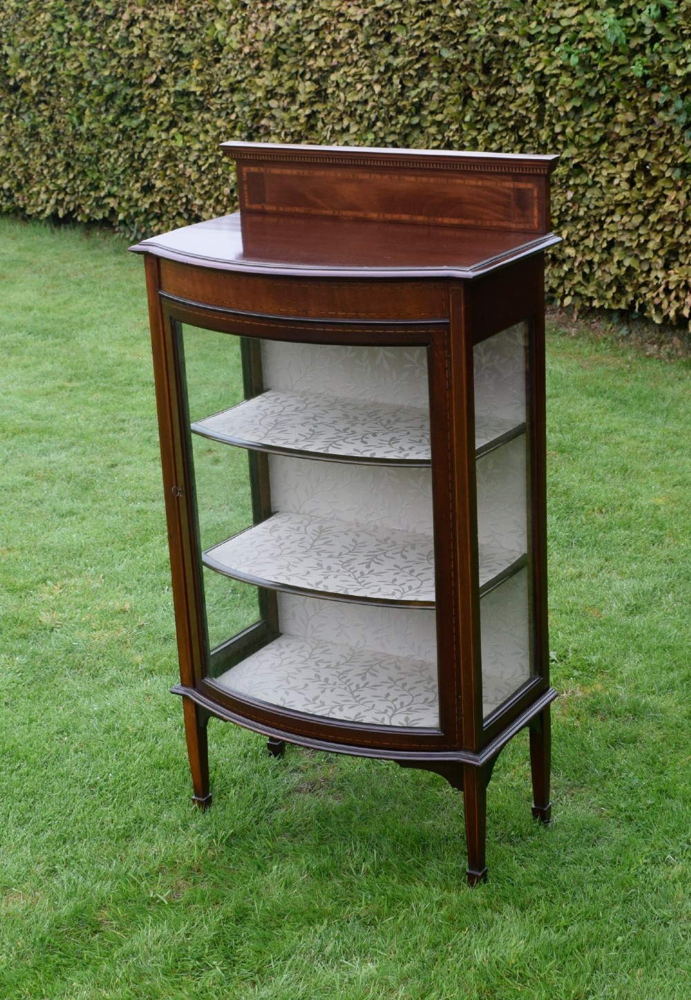 An Edwardian mahogany bowfront display cabinet featuring curved glass doors, satinwood string inlay, lined interior shelves, and tapered square legs. View XV