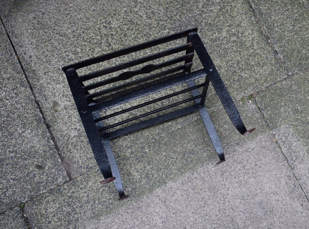 A black wrought iron fireplace footman featuring a slatted top with a central decorative cutout handle, resting on four curved legs with flat circular feet. View VII