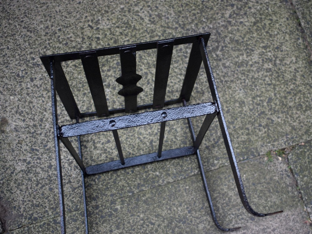 A black wrought iron fireplace footman featuring a slatted top with a central decorative cutout handle, resting on four curved legs with flat circular feet. View VIII