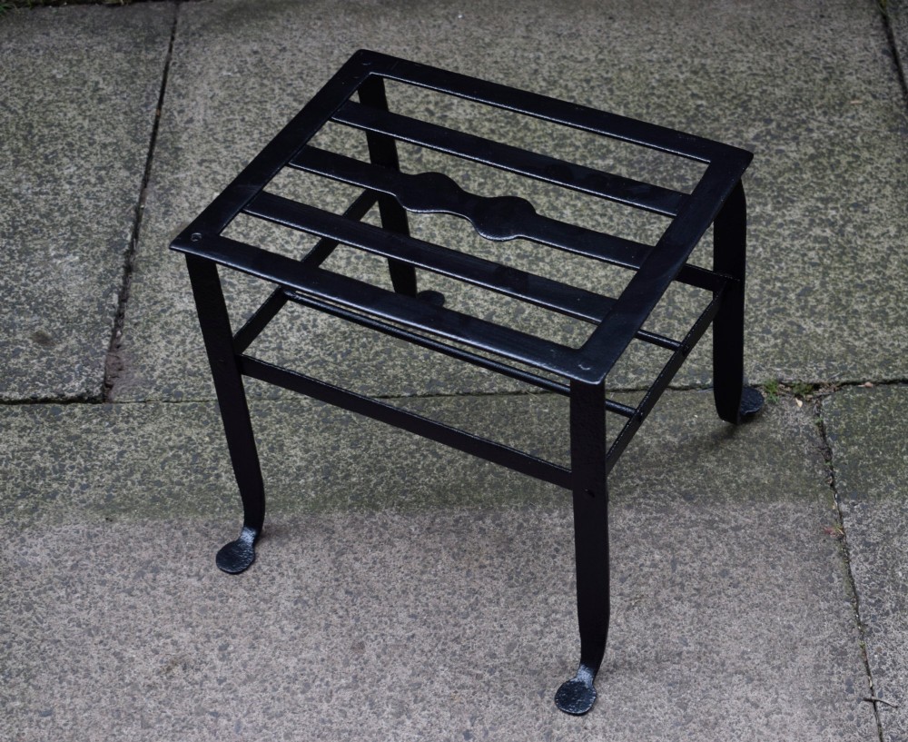 A black wrought iron fireplace footman featuring a slatted top with a central decorative cutout handle, resting on four curved legs with flat circular feet. View X
